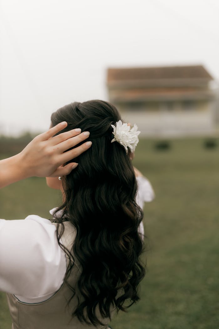 Woman with long dark hair and flower accessory outdoors, gently touching her hair.